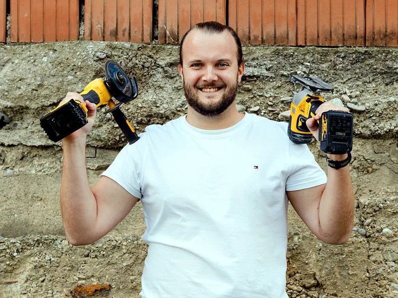 Christoph Harrison vor einer Ziegelmauer, hält lachend zwei Akku-Werkzeuge hoch